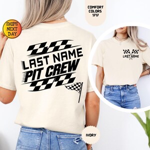 May include: A white t-shirt with a black checkered flag design and the text "Last Name Pit Crew". The shirt is shown on a model wearing blue jeans. The shirt is available in ivory.
