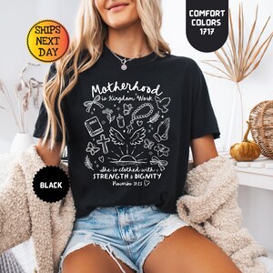 May include: Black t-shirt with white graphic text and illustrations. The text reads "Motherhood is Kingdom Work" and "She is clothed with strength & dignity". The shirt also features a cross, wings, and butterflies.