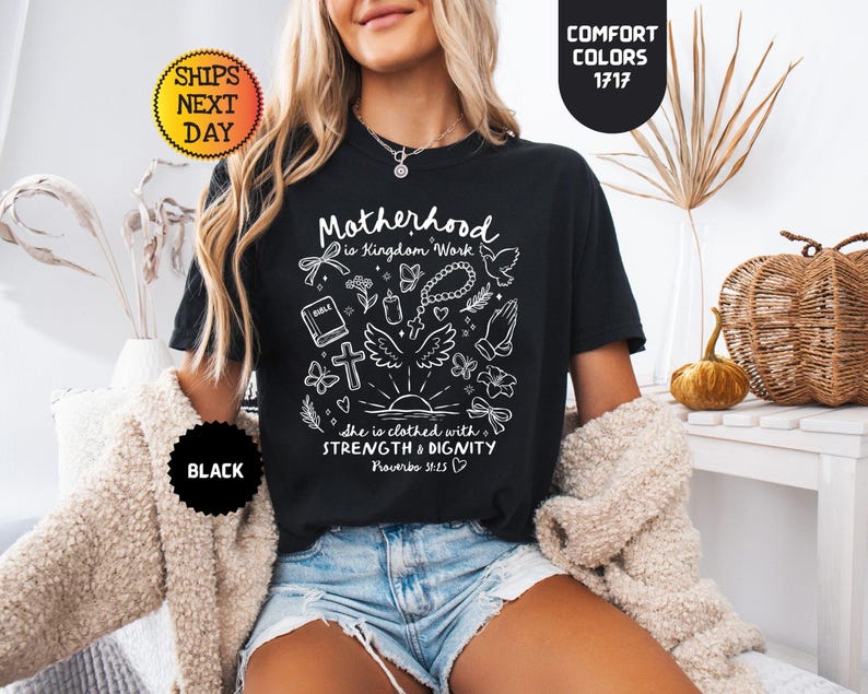 May include: Black t-shirt with white graphic text and illustrations. The text reads "Motherhood is Kingdom Work" and "She is clothed with strength & dignity". The shirt also features a cross, wings, and butterflies.