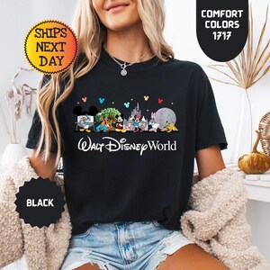 May include: Black t-shirt featuring a colorful graphic of Disney characters and landmarks, with the text "Walt Disney World" in white. The shirt includes the text "Comfort Colors 1717" and "Ships Next Day".