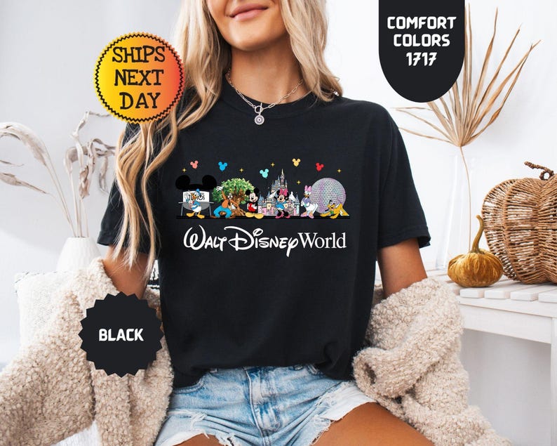 May include: Black t-shirt featuring a colorful graphic of Disney characters and landmarks, with the text "Walt Disney World" in white. The shirt includes the text "Comfort Colors 1717" and "Ships Next Day".