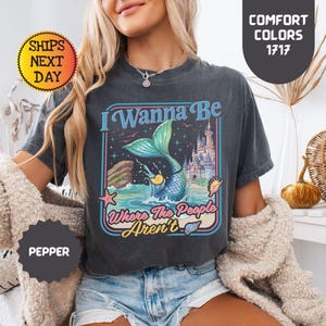 May include: A charcoal gray short-sleeve t-shirt featuring a graphic print of a mermaid tail splashing in the ocean, a castle, and the text I Wanna Be Where The People Aren't. The shirt has a crew neckline and a vintage-inspired washed finish.