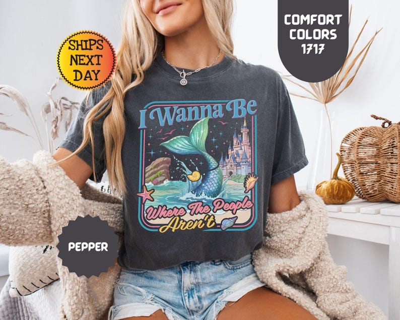 May include: A charcoal gray short-sleeve t-shirt featuring a graphic print of a mermaid tail splashing in the ocean, a castle, and the text I Wanna Be Where The People Aren't. The shirt has a crew neckline and a vintage-inspired washed finish.