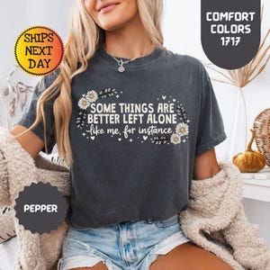 May include: Dark grey t-shirt with the text "SOME THINGS ARE BETTER LEFT ALONE like me, for instance" in white. The text is surrounded by floral and leaf designs. The shirt is a comfort color and the color is pepper.