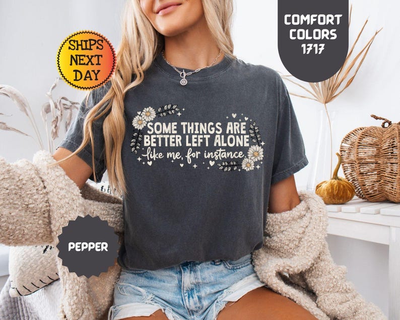May include: Dark grey t-shirt with the text "SOME THINGS ARE BETTER LEFT ALONE like me, for instance" in white. The text is surrounded by floral and leaf designs. The shirt is a comfort color and the color is pepper.