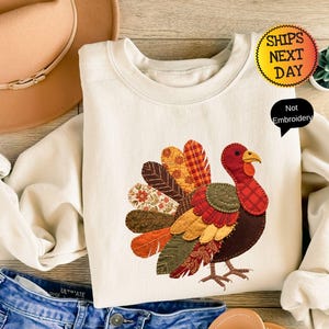 May include: A cream-colored crewneck sweatshirt featuring a central graphic of a turkey with a multi-patterned, colorful tail. The turkey design uses various fabric-like textures in shades of orange, red, brown, and green. A black speech bubble reads Not Embroidery.