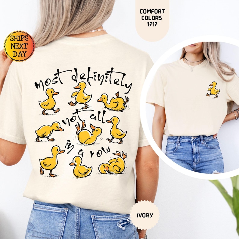 May include: Ivory-colored t-shirt featuring a playful design of yellow ducklings with the text "most definitely not all in a row." A small duckling graphic is on the front. The shirt is a Comfort Colors 1717.