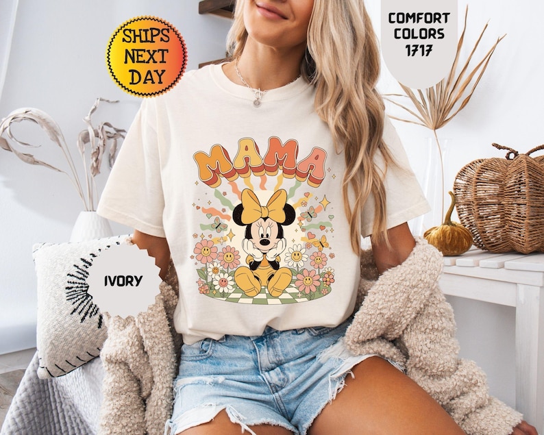 May include: Ivory-colored t-shirt featuring the word "MAMA" in retro orange and pink lettering, with a Minnie Mouse graphic. The design includes flowers, butterflies, and a yellow bow. The shirt is part of the Comfort Colors 1717 collection.