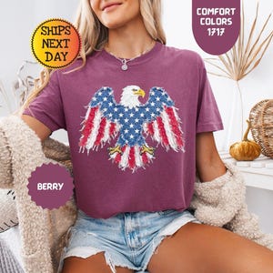 May include: Berry-colored t-shirt featuring an American flag eagle design. The eagle's wings are adorned with the stars and stripes. The shirt has the text "Comfort Colors 1717" and "Ships Next Day".