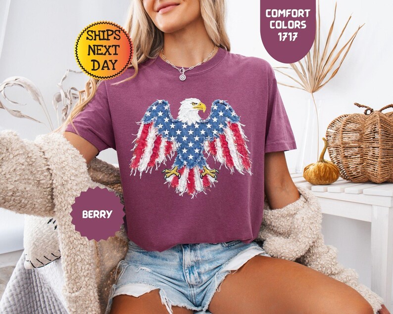 May include: Berry-colored t-shirt featuring an American flag eagle design. The eagle's wings are adorned with the stars and stripes. The shirt has the text "Comfort Colors 1717" and "Ships Next Day".