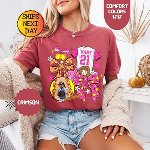 May include: A crimson short-sleeve t-shirt featuring a graphic print of a softball, a baseball bat, a glove, a jersey with the text NAME 21, and large bows. The design includes yellow, pink, and white accents with star and flower motifs.