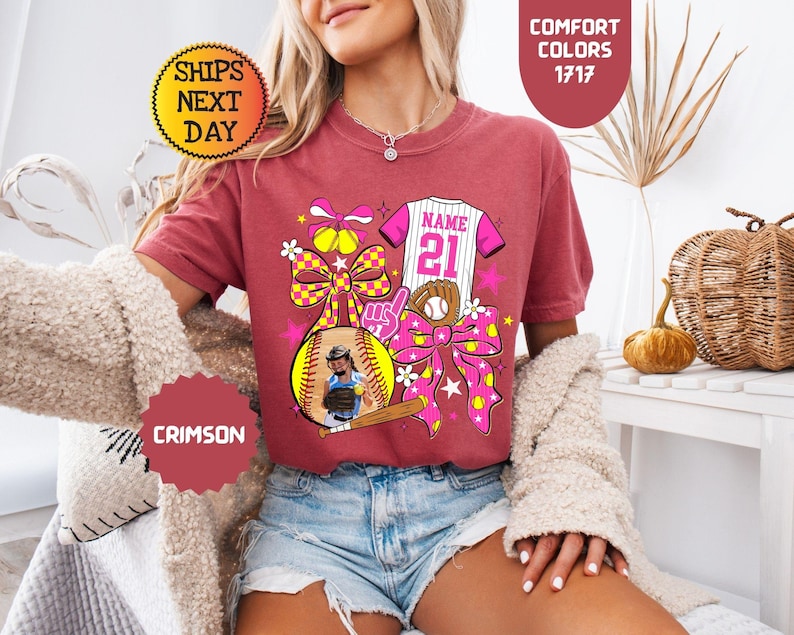 May include: A crimson short-sleeve t-shirt featuring a graphic print of a softball, a baseball bat, a glove, a jersey with the text NAME 21, and large bows. The design includes yellow, pink, and white accents with star and flower motifs.