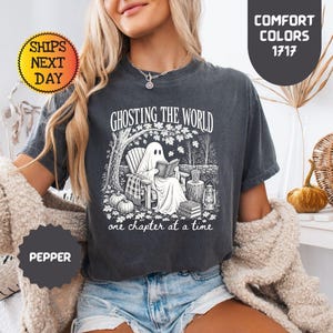 May include: A charcoal gray short-sleeve t-shirt featuring a white graphic print of a ghost sitting in a chair reading a book under a tree. The text reads GHOSTING THE WORLD one chapter at a time. The shirt has a crew neckline and a relaxed fit.