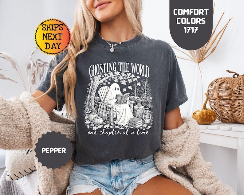 May include: A charcoal gray short-sleeve t-shirt featuring a white graphic print of a ghost sitting in a chair reading a book under a tree. The text reads GHOSTING THE WORLD one chapter at a time. The shirt has a crew neckline and a relaxed fit.