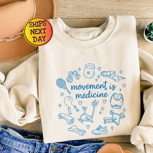 Könnte beinhalten: Ein cremefarbenes Sweatshirt mit Rundhalsausschnitt und einem blauen Grafikdruck verschiedener Fitnessgeräte, darunter eine Kettlebell, ein Tennisschläger und ein Heimtrainer, mit dem Text "movement is medicine" und kleinen Herz- und Sternsymbolen.