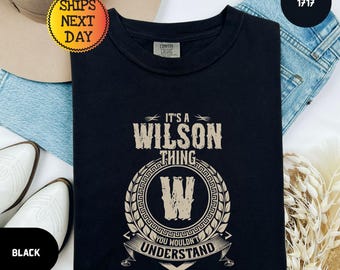 May include: A black short-sleeved t-shirt featuring a beige graphic print that reads IT'S A WILSON THING YOU WOULDN'T UNDERSTAND with a large letter W centred inside a circular laurel wreath design. The shirt has a crew neckline and a tag.