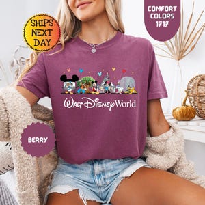 May include: Berry-colored t-shirt featuring a Walt Disney World graphic with various Disney characters and park icons. The text "Walt Disney World" is printed in white script below the graphic. The shirt has short sleeves and a crew neck.