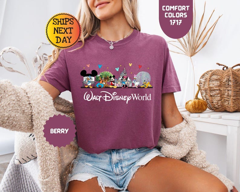 May include: Berry-colored t-shirt featuring a Walt Disney World graphic with various Disney characters and park icons. The text "Walt Disney World" is printed in white script below the graphic. The shirt has short sleeves and a crew neck.