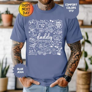 Könnte beinhalten: Blaues Jeans-T-Shirt mit einem weißen Grafikdesign mit dem Wort "daddy" und verschiedenen Symbolen wie einem Auto, Werkzeugen und Sportgeräten. Das Shirt enthält auch den Text "Fixer of Everything" und "Grill Master (Sometimes)."