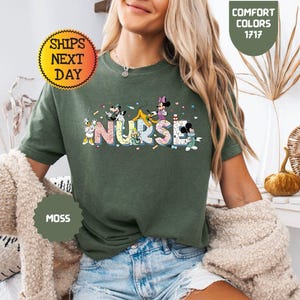 May include: Moss green t-shirt with the word "NURSE" in colorful, cartoon-style lettering. The letters are decorated with various cartoon characters. The shirt also has the text "COMFORT COLORS 1717" and "SHIPS NEXT DAY" on the shirt.