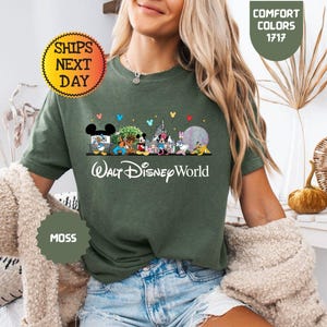 May include: Moss green t-shirt featuring a colorful graphic of Disney characters and landmarks, including Mickey Mouse, Minnie Mouse, and Cinderella's castle. The text "Walt Disney World" is printed below the graphic. The shirt also has the text "Comfort Colors 1717" and "Ships Next Day".