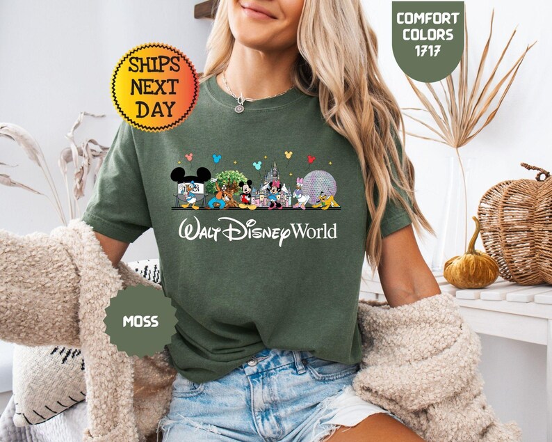 May include: Moss green t-shirt featuring a colorful graphic of Disney characters and landmarks, including Mickey Mouse, Minnie Mouse, and Cinderella's castle. The text "Walt Disney World" is printed below the graphic. The shirt also has the text "Comfort Colors 1717" and "Ships Next Day".