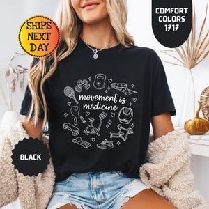 May include: Black t-shirt with the phrase "movement is medicine" surrounded by white line art of exercise equipment. The shirt has a crew neck and short sleeves. The image also includes text that says "Comfort Colors 1717" and "Ships Next Day".