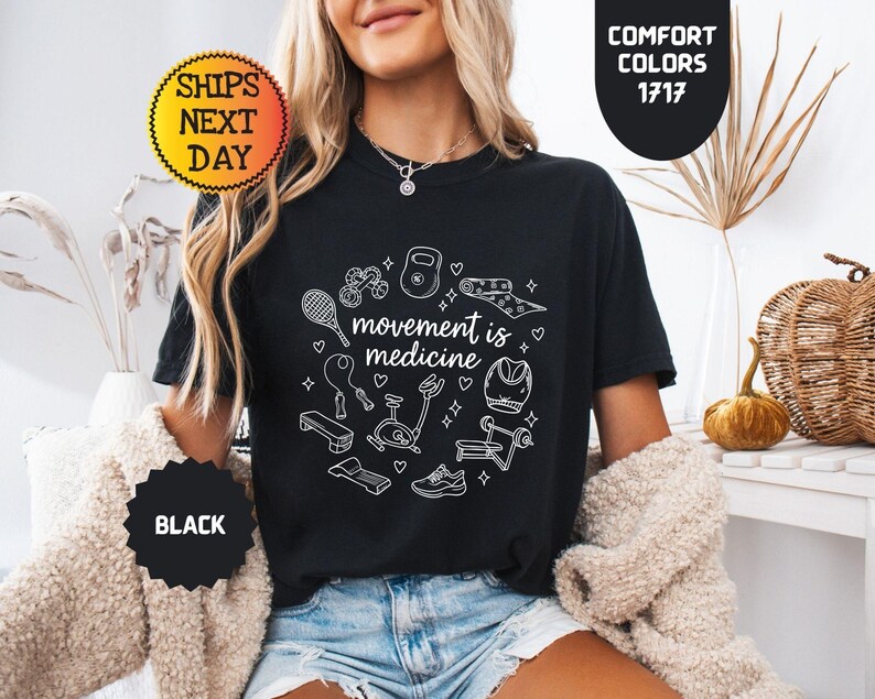 May include: Black t-shirt with the phrase "movement is medicine" surrounded by white line art of exercise equipment. The shirt has a crew neck and short sleeves. The image also includes text that says "Comfort Colors 1717" and "Ships Next Day".