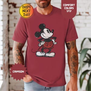 May include: Crimson t-shirt with a distressed Mickey Mouse graphic. The cartoon character is in black, white, and red. The shirt has short sleeves and a crew neck. The text "Comfort Colors 1717" is visible. The shirt is a comfort color.