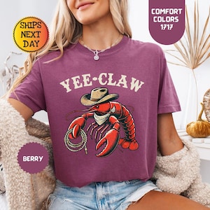 以下が含まれることがあります： ベリー色のTシャツ。カウボーイハットとロープを身につけた赤いロブスターのグラフィックが特徴で、上部に「Yee-Claw」の文字。Comfort Colorsコレクションの一部です。ゆったりとしたフィット感のシャツです。