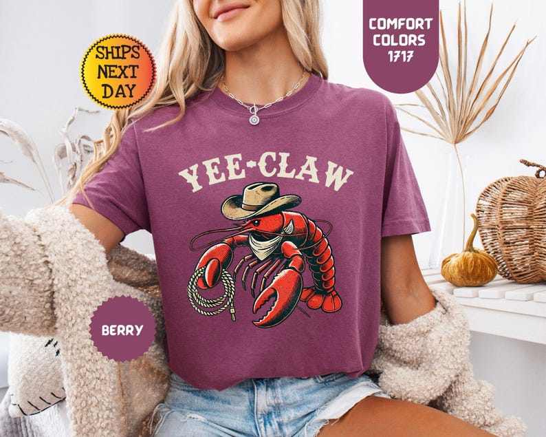 以下が含まれることがあります： ベリー色のTシャツ。カウボーイハットとロープを身につけた赤いロブスターのグラフィックが特徴で、上部に「Yee-Claw」の文字。Comfort Colorsコレクションの一部です。ゆったりとしたフィット感のシャツです。