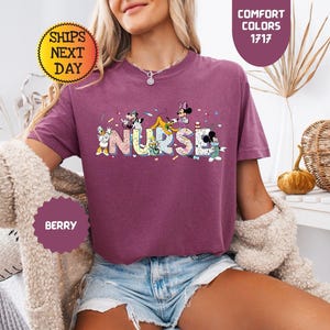 May include: Berry-colored t-shirt with the word "NURSE" in a colorful, playful font, featuring cartoon characters. The shirt has short sleeves and a crew neck. The image also includes text that says "Comfort Colors 1717" and "Ships Next Day".