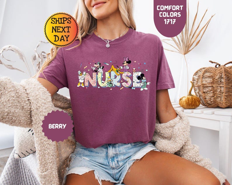 May include: Berry-colored t-shirt with the word "NURSE" in a colorful, playful font, featuring cartoon characters. The shirt has short sleeves and a crew neck. The image also includes text that says "Comfort Colors 1717" and "Ships Next Day".