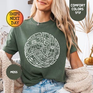 May include: A moss green t-shirt featuring a white volleyball design with floral patterns. The shirt has the text "Comfort Colors 1717" and "Moss". The image also includes the text "Ships Next Day".