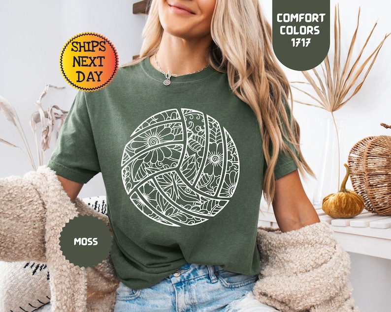 May include: A moss green t-shirt featuring a white volleyball design with floral patterns. The shirt has the text "Comfort Colors 1717" and "Moss". The image also includes the text "Ships Next Day".