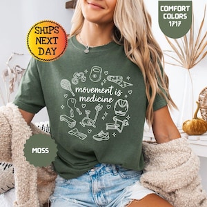 May include: A moss green t-shirt with the phrase "movement is medicine" in a white circular design. The design includes illustrations of exercise equipment. The shirt is a comfort color, and the image includes a "Ships Next Day" sticker.