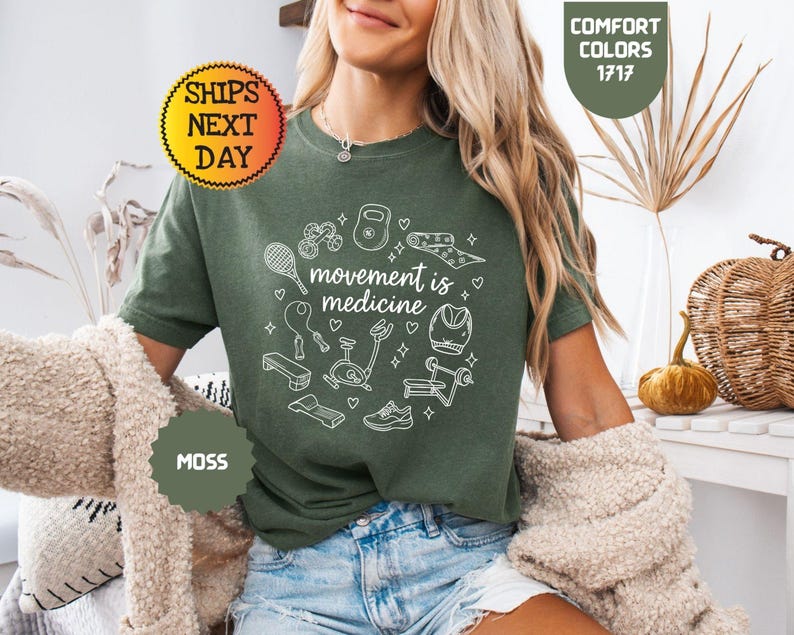 May include: A moss green t-shirt with the phrase "movement is medicine" in a white circular design. The design includes illustrations of exercise equipment. The shirt is a comfort color, and the image includes a "Ships Next Day" sticker.