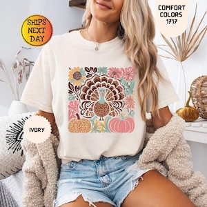 以下が含まれることがあります： アイボリーの半袖Tシャツ。中央にはヒョウ柄のディテールが施された七面鳥のグラフィックプリントがあり、その周囲をオレンジ、ピンク、ティールカラーのカラフルな花、葉、2つのカボチャが囲んでいます。クルーネック仕様です。