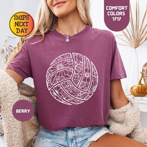 May include: Berry-colored t-shirt featuring a white, detailed volleyball design. The shirt has short sleeves and a crew neck. The text "Comfort Colors 1717" is printed on the top right. The text "Ships Next Day" is printed on the top left. The text "Berry" is printed on the bottom left.