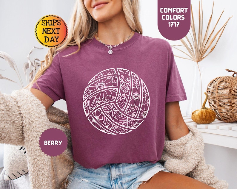 May include: Berry-colored t-shirt featuring a white, detailed volleyball design. The shirt has short sleeves and a crew neck. The text "Comfort Colors 1717" is printed on the top right. The text "Ships Next Day" is printed on the top left. The text "Berry" is printed on the bottom left.