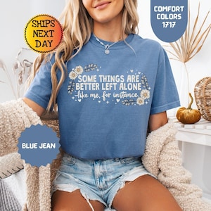 May include: A blue jean colored t-shirt with the text "SOME THINGS ARE BETTER LEFT ALONE like me, for instance" in white. The shirt has a floral design and is part of the Comfort Colors 1717 collection.
