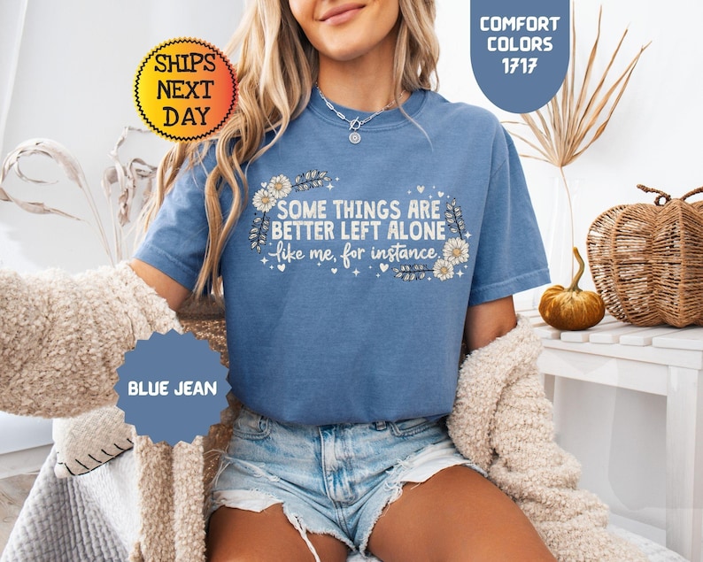 May include: A blue jean colored t-shirt with the text "SOME THINGS ARE BETTER LEFT ALONE like me, for instance" in white. The shirt has a floral design and is part of the Comfort Colors 1717 collection.