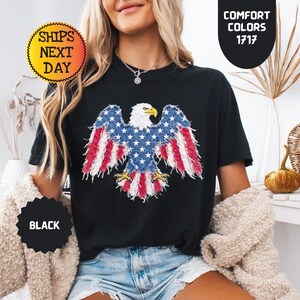 May include: Black t-shirt featuring a patriotic design of an American bald eagle with the US flag incorporated into the wings. The shirt has the text "Comfort Colors 1717" and "Ships Next Day".