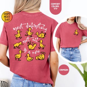 May include: Crimson-colored t-shirt with a playful design of yellow ducklings and the phrase "most definitely not all in a row." A small duckling graphic is on the front. The shirt has a round neck and short sleeves.
