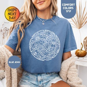 May include: Blue jean colored t-shirt with a white volleyball design. The volleyball is decorated with floral and geometric patterns. The shirt has the text "Comfort Colors 1717" and "Blue Jean" on the image.