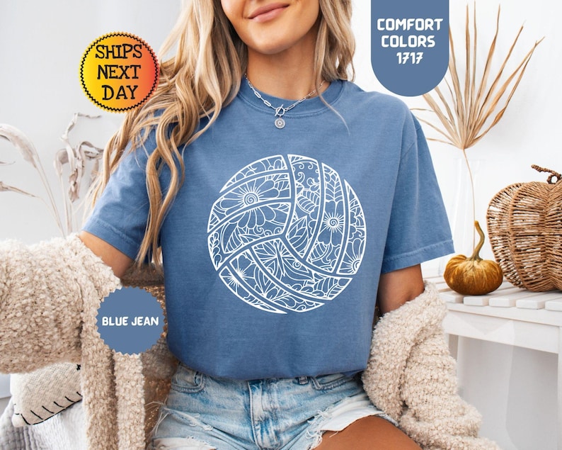 May include: Blue jean colored t-shirt with a white volleyball design. The volleyball is decorated with floral and geometric patterns. The shirt has the text "Comfort Colors 1717" and "Blue Jean" on the image.