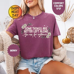 May include: Berry-colored t-shirt with the text "SOME THINGS ARE BETTER LEFT ALONE like me, for instance." The shirt has a floral design and is part of the Comfort Colors collection.