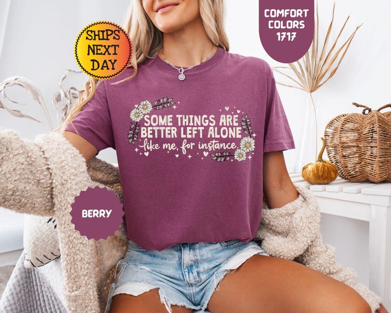 May include: Berry-colored t-shirt with the text "SOME THINGS ARE BETTER LEFT ALONE like me, for instance." The shirt has a floral design and is part of the Comfort Colors collection.