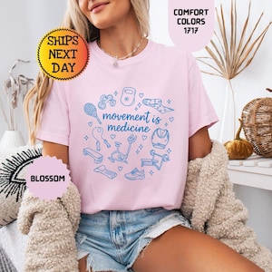 May include: A light pink t-shirt with the phrase "movement is medicine" in a blue circular design with fitness equipment illustrations. The shirt is worn with denim shorts and a cream-colored sweater.