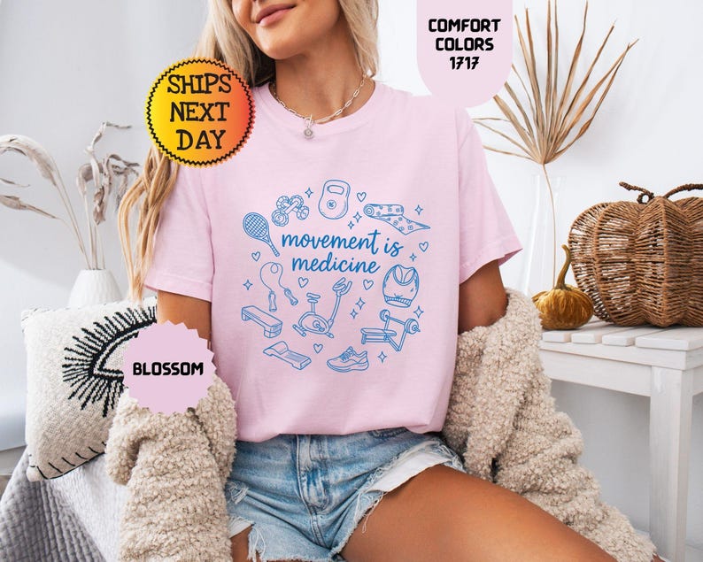 May include: A light pink t-shirt with the phrase "movement is medicine" in a blue circular design with fitness equipment illustrations. The shirt is worn with denim shorts and a cream-colored sweater.